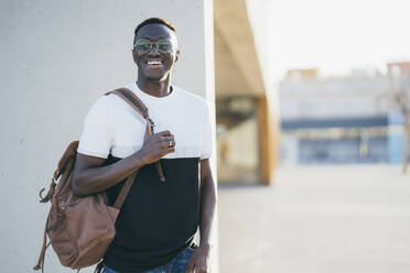 Smiling man with backpack looking away while standing by wall - MPPF01704