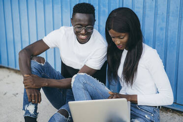 Woman using laptop while sitting with boyfriend near blue wall - MPPF01714