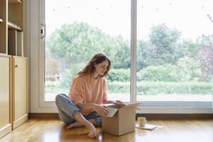 Schöne Frau beim Öffnen eines Kartons, während sie zu Hause sitzt - AFVF08703