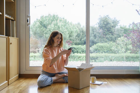 Lächelnde Frau, die einen Karton mit ihrem Smartphone fotografiert, während sie zu Hause sitzt - AFVF08704