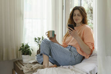 Beautiful woman with coffee cup using smart phone while sitting on mattress at home - AFVF08708