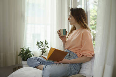 Nachdenkliche Frau mit Buch, die wegschaut, während sie zu Hause auf einer Matratze sitzt - AFVF08709
