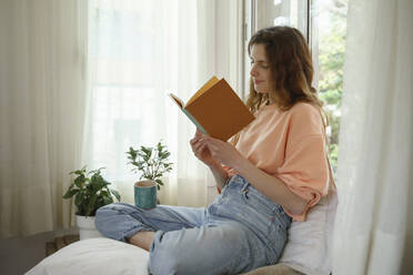 Woman reading book while sitting on mattress at home - AFVF08710