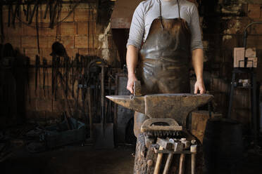 Männlicher Schmied stehend mit Hammer am Amboss in der Werkstatt - AODF00480