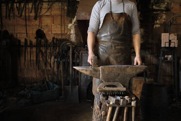 Male blacksmith standing with hammer by anvil at shop - AODF00480