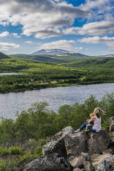 Mother and baby son admiring view of Karasjohka river while sitting on top of boulder - RUNF04356