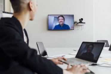 Geschäftsfrau, die an einer Besprechung mit einem Kollegen per Videoanruf über Laptop und Fernseher im Büro teilnimmt - MEUF02540