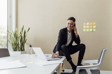 Fröhlicher Unternehmer, der mit dem Handy telefoniert, während er einen Laptop im Büro benutzt - MEUF02546