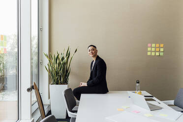 Lächelnde Geschäftsfrau sitzt auf dem Schreibtisch an der Wand im Büro - MEUF02553