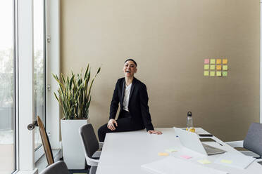 Cheerful businesswoman sitting on desk by window in office - MEUF02554