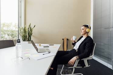 Geschäftsfrau mit Kopfhörern und Kaffeetasse vor einem Laptop im Büro - MEUF02583