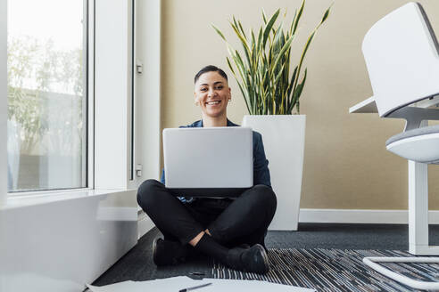 Lächelnde Geschäftsfrau mit Laptop auf dem Schoß sitzt auf dem Boden im Büro - MEUF02603