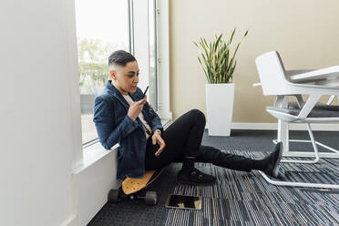 Berufstätige Frau sendet Sprachnachrichten über ihr Mobiltelefon, während sie auf einem Skateboard im Büro sitzt - MEUF02606