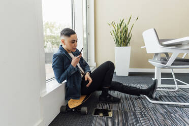 Female professional sending voice mail through mobile phone while sitting on skateboard in office - MEUF02606