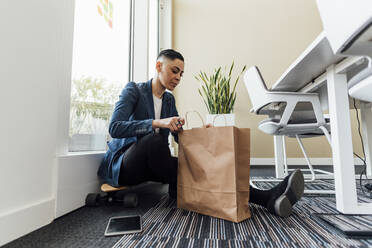 Geschäftsfrau schaut auf Papiertüte, während sie im Büro sitzt - MEUF02610