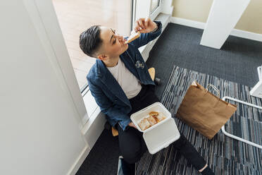 Unternehmerin beim Essen, während sie im Büro am Fenster sitzt - MEUF02615