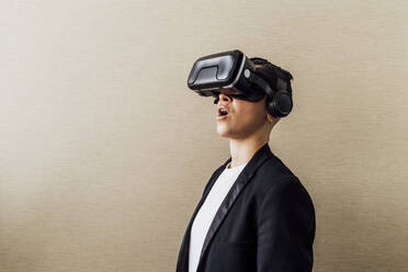 Geschäftsfrau steht mit offenem Mund, während sie ein Virtual-Reality-Headset an einer Wand im Büro benutzt - MEUF02651