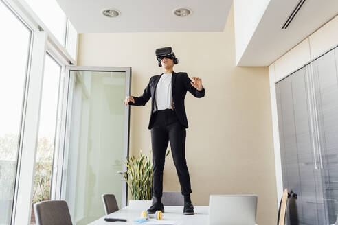 Junge Geschäftsfrau, die einen Virtual-Reality-Simulator benutzt, während sie auf einem Schreibtisch im Büro steht - MEUF02655