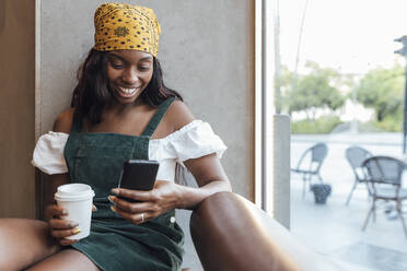 Frau mit Kopftuch hält Kaffeetasse und benutzt Smartphone im Café - JRVF00507