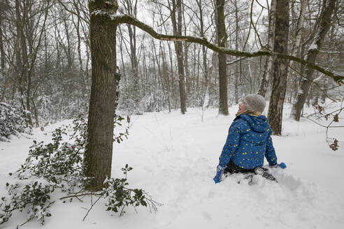 Junge schaut weg, während er im Winter in der Nähe eines Baumes im Schnee kniet - FVDF00004