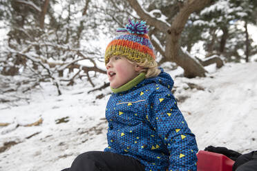 Neugieriger Junge schaut weg, während er im Winter auf dem Schnee sitzt - FVDF00012