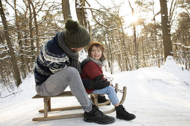 Father looking at son while sitting on sled during winter - FVDF00039