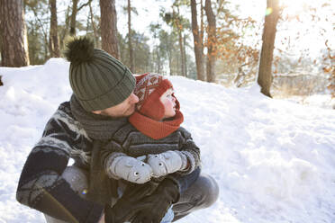 Vater mit Strickmütze, der seinen Sohn umarmt, während er im Winter im Schnee hockt - FVDF00072