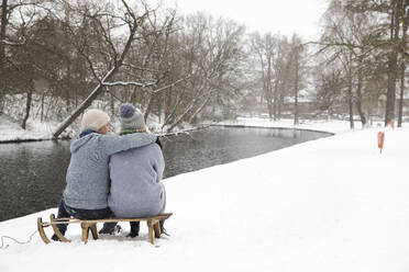 Paar umarmt, während auf Schlitten in der Nähe von See im Winter sitzen - FVDF00088