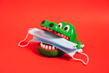 Plastic crocodile teeth toy with medical mask in teeth placed on red background in studio - ADSF23457