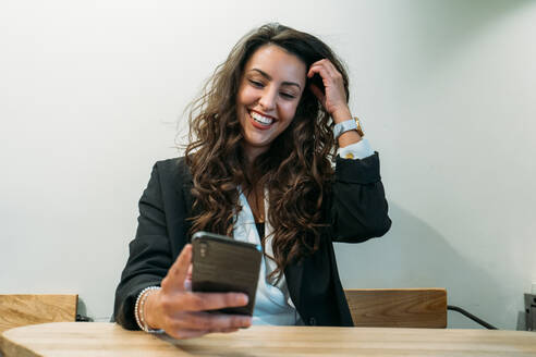 Fröhliche Unternehmerin im schicken Anzug, die am Tisch sitzt und ihr Smartphone benutzt, während sie aus der Ferne in einem Café arbeitet und auf eine Bestellung wartet - ADSF23523