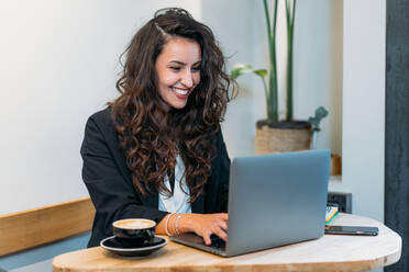 Charmante Unternehmerin am Tisch sitzend mit Kaffee in einer Tasse und einem Laptop während der Arbeit an einem Projekt aus der Ferne in einem Café - ADSF23527