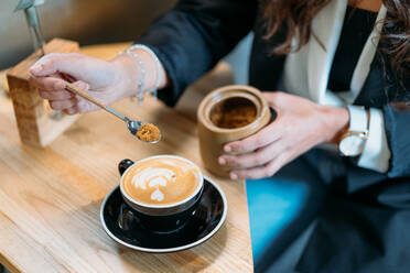 Von oben der Ernte anonyme weibliche Unternehmerin in stilvollem Outfit Zugabe Löffel braunen Zucker in Tasse mit Cappuccino während der Kaffeepause im Café - ADSF23528