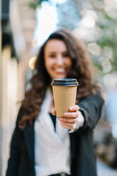 Cheerful blurred female entrepreneur standing on street and reaching out hand with takeaway coffee in paper cup towards camera - ADSF23529
