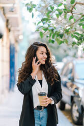 Positive Unternehmerin in lässigem Stil und mit Kaffee zum Mitnehmen im Pappbecher, die auf der Straße steht und mit ihrem Smartphone spricht - ADSF23530