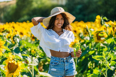 Fröhliche ethnische Frau mit Hut steht mit erhobenen Armen in einem blühenden Sonnenblumenfeld und schaut in die Kamera - ADSF23544