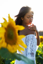 Thoughtful ethnic female standing in booming sunflower field and looking away - ADSF23550
