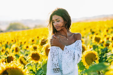 Thoughtful ethnic female standing in booming sunflower field and looking away - ADSF23551