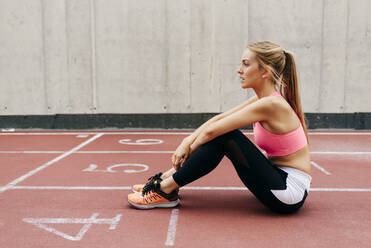 Seitenansicht einer blonden jungen Sportlerin, die auf dem Boden sitzt und sich auf einem Rollfeld ausruht und wegschaut - ADSF23559