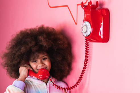 Lächelndes ethnisches Mädchen mit Afro-Frisur, das in einem Studio ein altmodisches rotes Telefon benutzt und in die Kamera auf rosa Hintergrund schaut - ADSF23601
