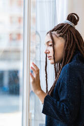 Pensive woman with dreadlocks looking out the window on a cloudy day - ADSF23603