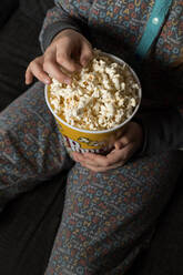 Von oben abgeschnittenes, nicht erkennbares Kind, das gemütlich auf dem Sofa mit einem Eimer Popcorn sitzt, während es isst und fernsieht - ADSF23618