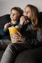 Junge staunende Geschwisterkinder sitzen bequem auf dem Sofa mit Popcorn-Eimer beim Essen und Fernsehen - ADSF23619