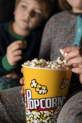 Cropped unscharf junge erstaunt Geschwister Kinder sitzen bequem auf dem Sofa mit Popcorn-Eimer beim Essen und Fernsehen - ADSF23620