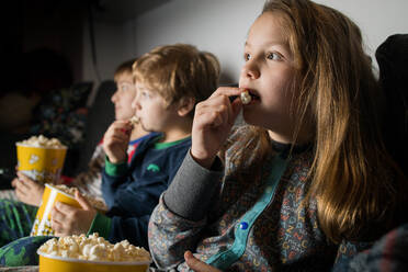 Junge staunende Geschwisterkinder sitzen bequem auf dem Sofa mit Popcorn-Eimer beim Essen und Fernsehen - ADSF23625