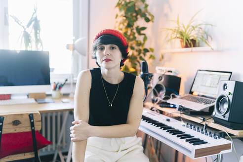 Female music composer wearing knit hat while sitting by electric piano in studio - DGOF02179