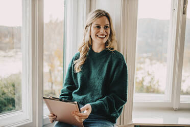 Blondhaarige Frau in Sweatshirt sitzt mit Tablet am Fenster im Büro - DAWF01970