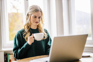 Geschäftsfrau hält Kaffeetasse und benutzt Laptop im Büro - DAWF01975