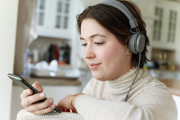 Schöne Frau trägt Kopfhörer, während sie zu Hause ein Mobiltelefon benutzt - EYAF01597