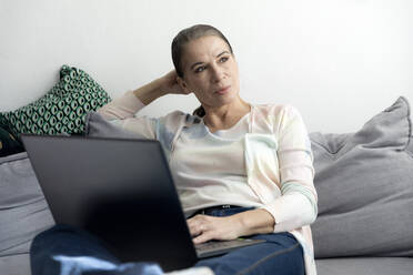 Nachdenkliche Frau sitzt mit Laptop auf dem Sofa zu Hause - FLLF00603