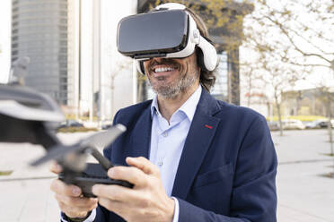 Mature male entrepreneur wearing virtual reality headset while holding drone in office park - JCCMF02034